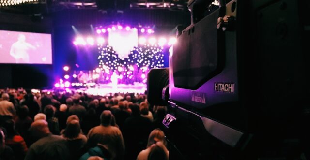 Large camera on tripod, pointed at purple stage and audience; coronavirus