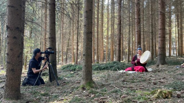 Woman sitting in the woods beating a drum in front of a camera